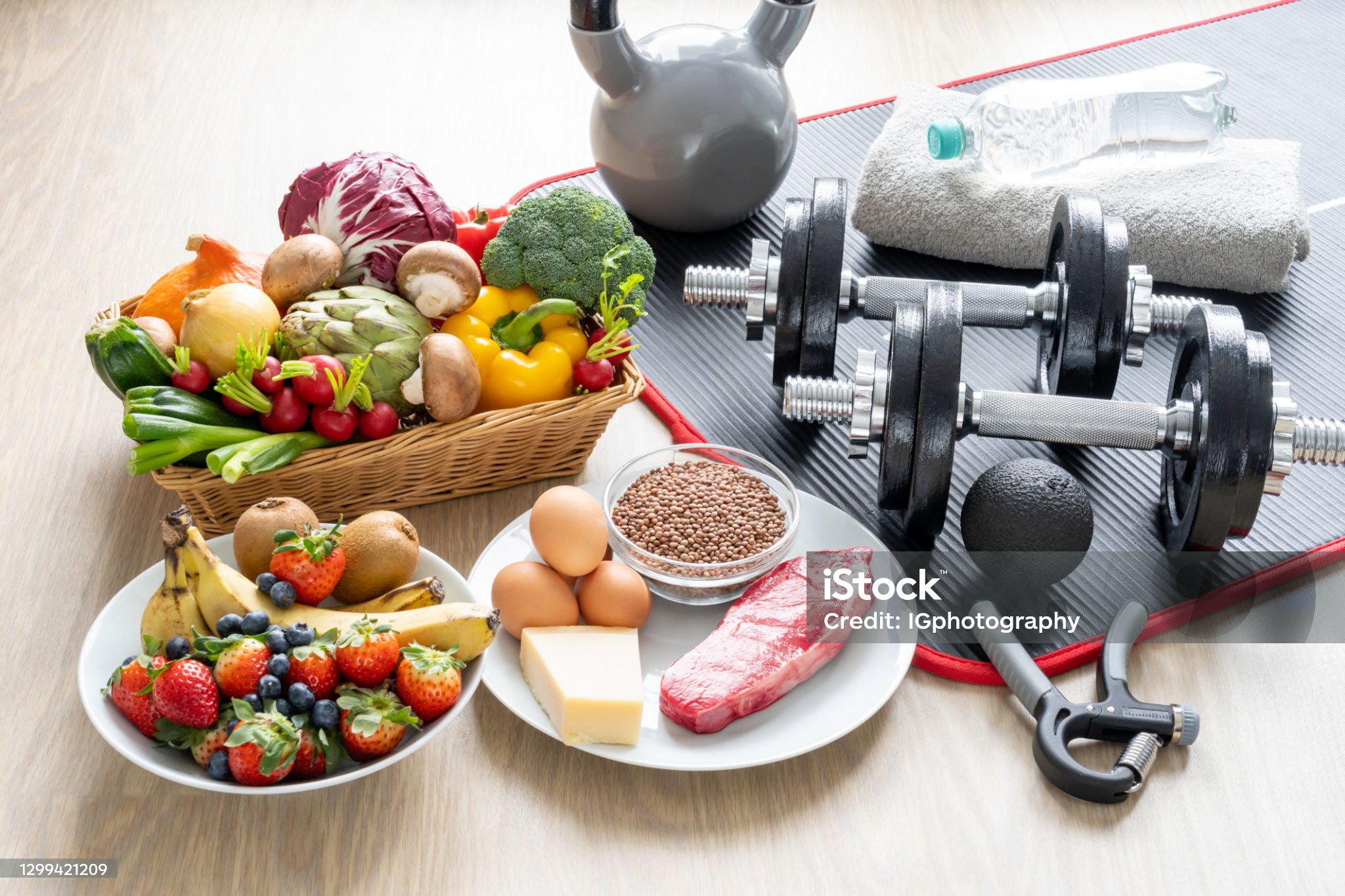 Healthy foods next to work out equipment. In the food basket there are fresh vegetables, including zucchinis, radishes, broccoli, artichokes, leaks, mushrooms, and bell peppers. There is also a fruit basked with kiwis, strawberries, blueberries, and bananas. There is also a plate with low carb healthy sources of protein, such a eggs, hard cheese, lentils and a fresh cut of meat (steak). There are also a pair of dumbbells, a grip strengthener, a kettlebell, a foam rolling ball, a yoga mat as well as a water bottle on top of a towel. This image illustrates the concept of healthy eating and exercising which is the best way to stay healthy, loose weight and live a long and happy life.